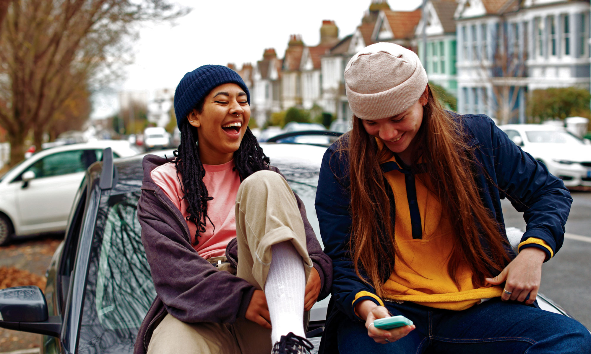 Two young adult women sitting at the hood of a car laughing
