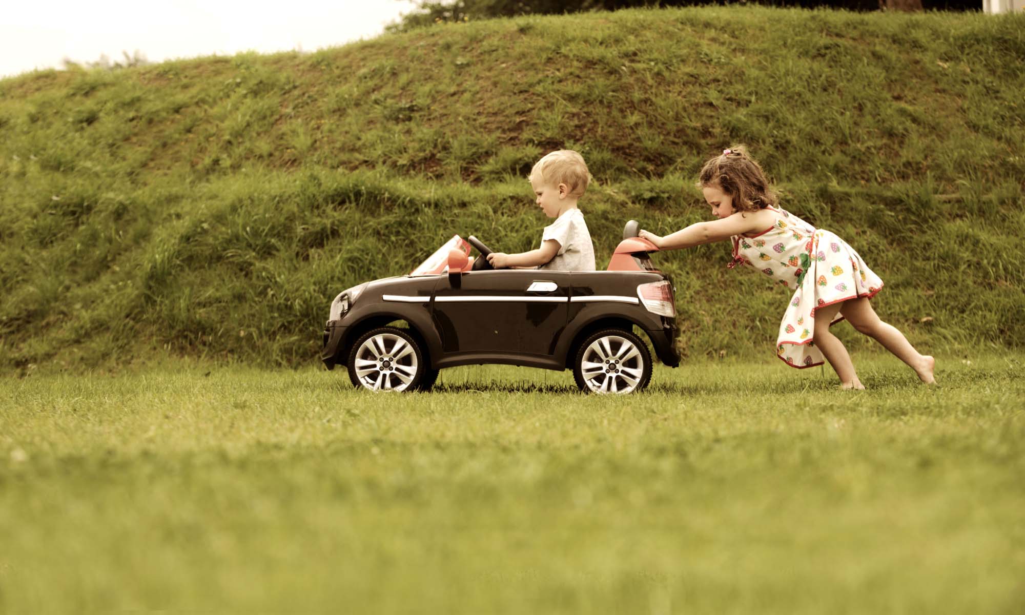 Child sitting in a toy car with another child pushing 