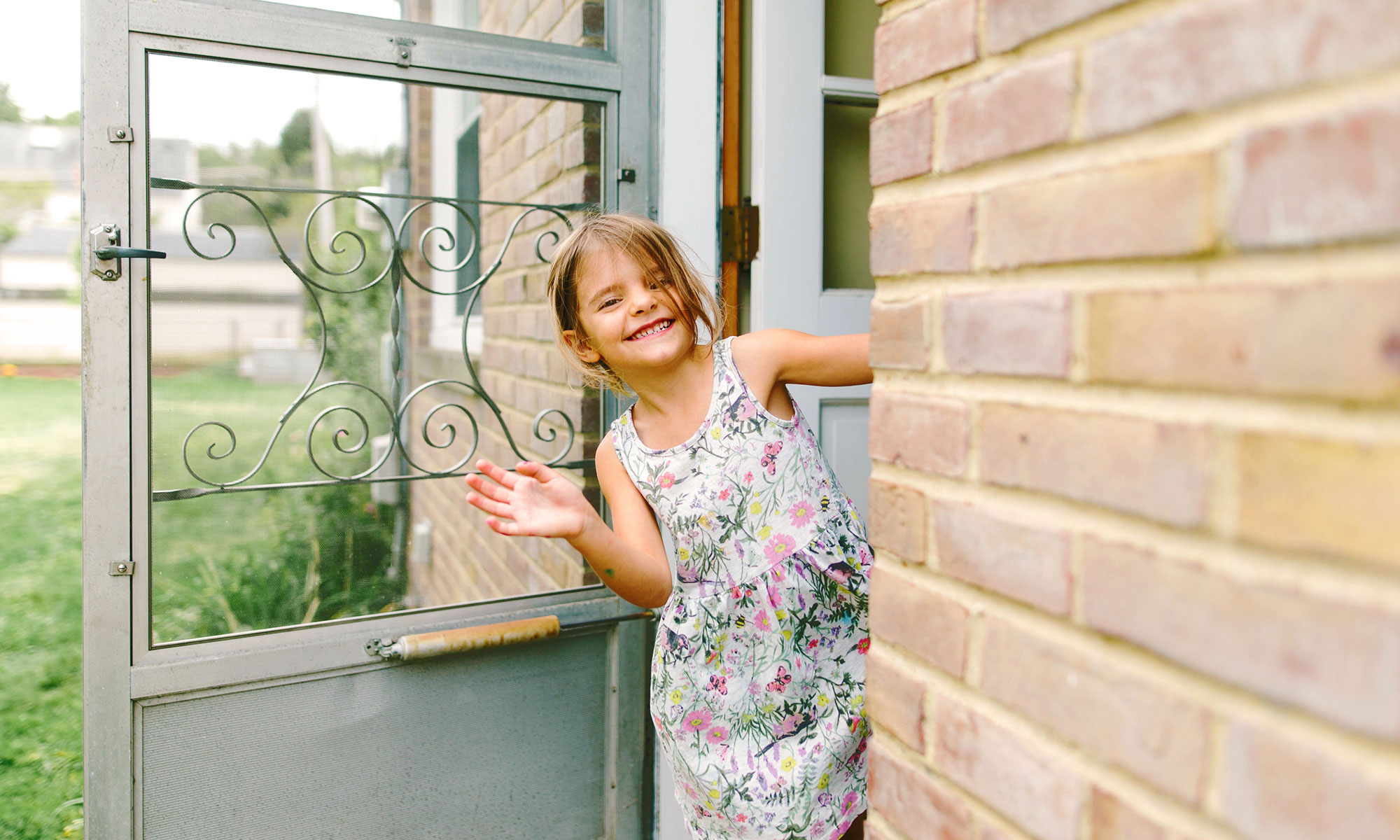 Small girl waving while leaning out her front door of her brick house