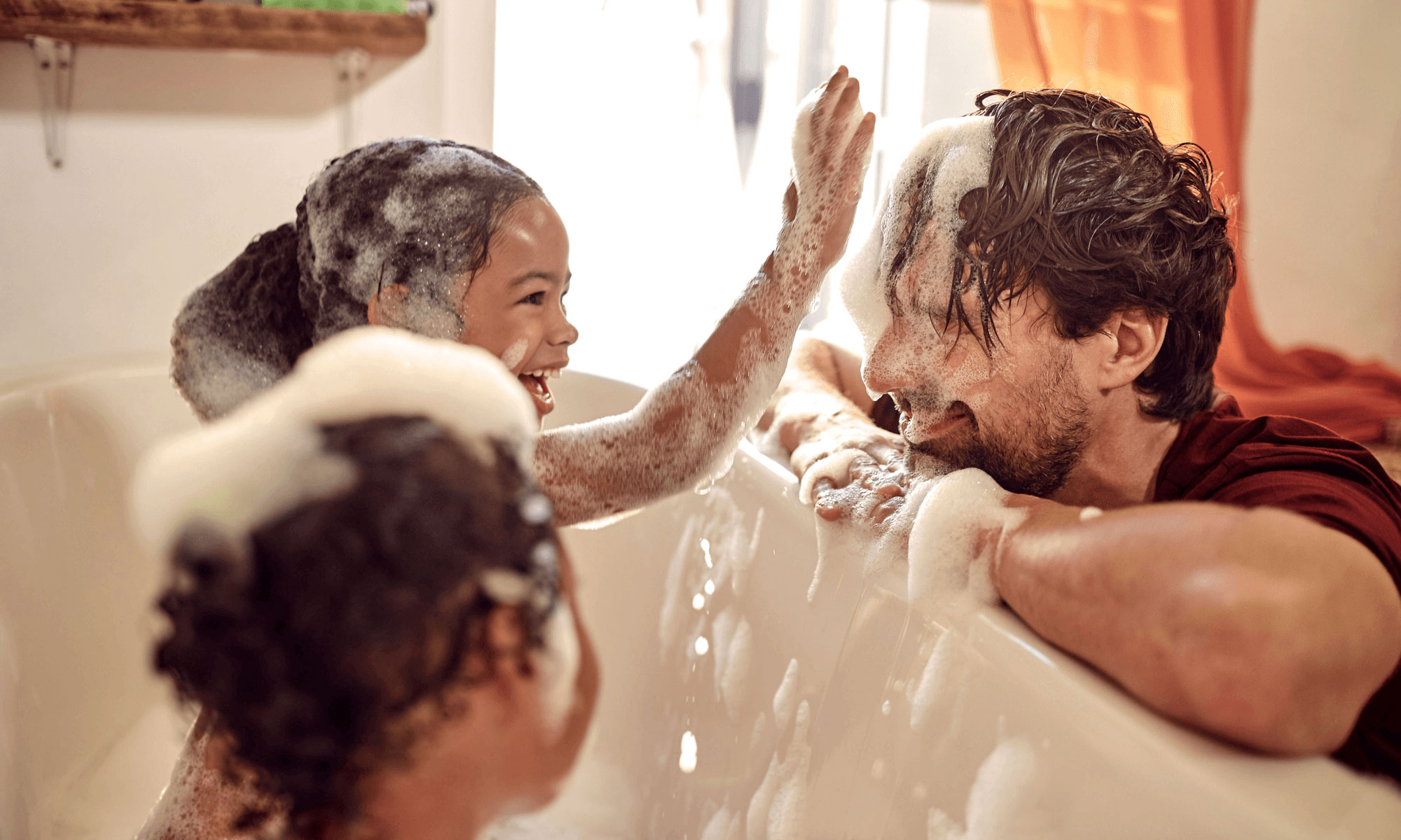 Father covered in suds while playing with his daughters in the tub, they're laughing.