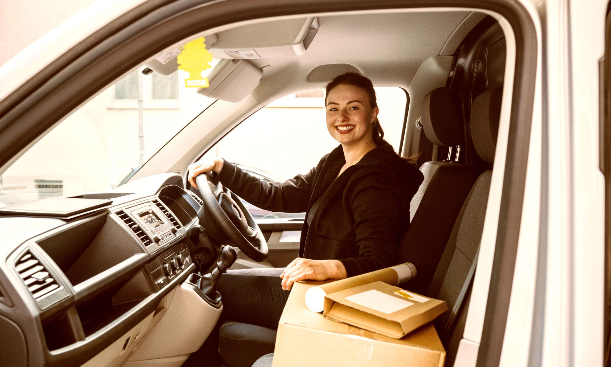Woman smiling while sitting in the driver's seat of a white van with some parcels in the foreground
